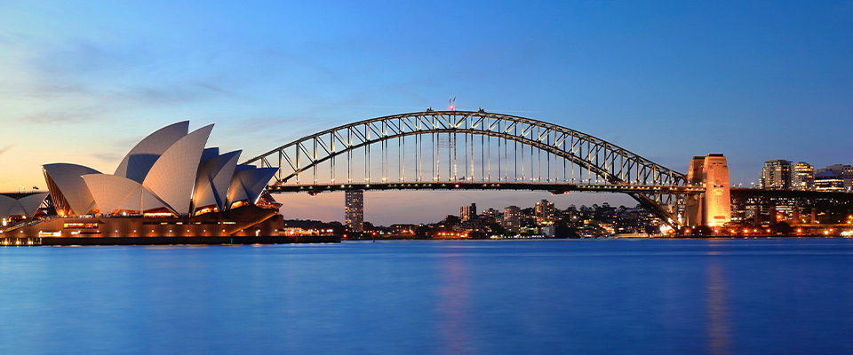 Vista panorâmica do Sydney Opera House e da Sydney Harbour Bridge ao entardecer. O céu exibe tons de azul e laranja típicos do pôr do sol. As luzes da cidade e dos edifícios estão acesas, refletindo na água calma do porto. À esquerda, o Sydney Opera House se destaca com seu design icônico em forma de velas brancas. Ao centro, a Sydney Harbour Bridge, uma ponte arqueada de aço, cruza a imagem. Ao fundo, arranha-céus iluminados compõem o horizonte da cidade de Sydney.