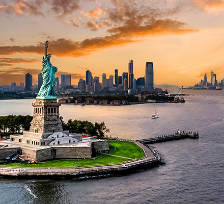 Imagem da Estátua da Liberdade em primeiro plano, situada na Liberty Island, cercada pelas águas do rio Hudson. Ao fundo, vê-se o horizonte da cidade de Nova York com diversos arranha-céus, incluindo o One World Trade Center à direita. O céu está parcialmente nublado, com tons alaranjados e rosados típicos do pôr do sol. Alguns barcos estão visíveis na água, compondo uma cena urbana e simbólica dos Estados Unidos.