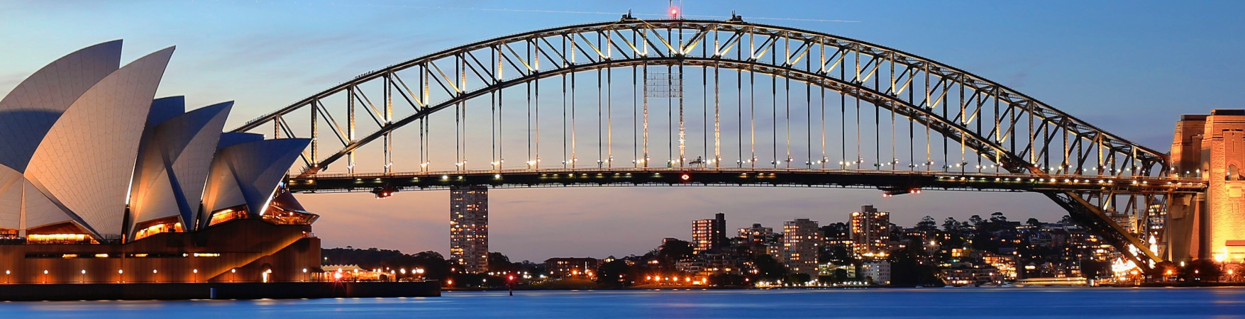 Vista panorâmica do Sydney Opera House e da Sydney Harbour Bridge ao entardecer. O céu exibe tons de azul e laranja típicos do pôr do sol. As luzes da cidade e dos edifícios estão acesas, refletindo na água calma do porto. À esquerda, o Sydney Opera House se destaca com seu design icônico em forma de velas brancas. Ao centro, a Sydney Harbour Bridge, uma ponte arqueada de aço, cruza a imagem. Ao fundo, arranha-céus iluminados compõem o horizonte da cidade de Sydney.