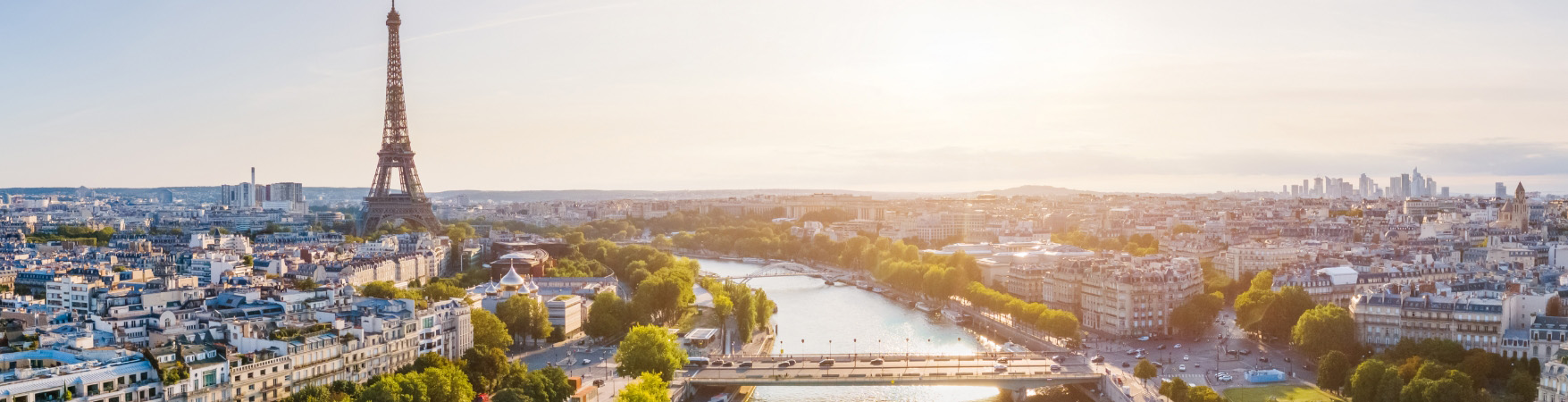 Vista panorâmica de Paris em um dia ensolarado. À esquerda, a Torre Eiffel se destaca no horizonte. O rio Sena atravessa o centro da imagem, com embarcações atracadas ao longo das margens. A cidade se estende dos dois lados do rio, com uma mistura de edifícios históricos e modernos. Árvores e vegetação verde acompanham as margens, trazendo contraste à paisagem urbana.