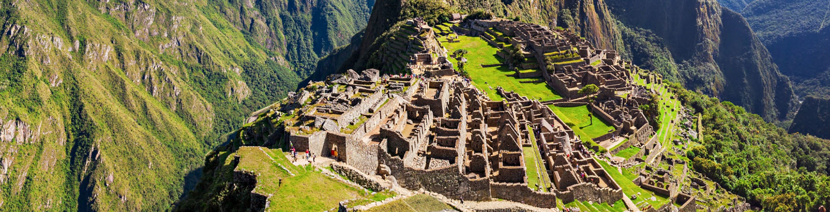 Vista panorâmica de Machu Picchu, antigo sítio arqueológico inca no Peru. A cidade está localizada no topo de uma montanha, com terraços verdes e ruínas de pedra bem preservadas. Ao fundo, montanhas altas cobertas por vegetação se destacam sob um céu azul parcialmente nublado. A imagem combina a grandiosidade das construções históricas com a beleza natural da paisagem montanhosa.