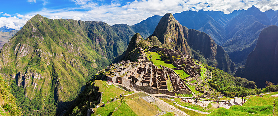 Vista panorâmica de Machu Picchu, antigo sítio arqueológico inca no Peru. A cidade está localizada no topo de uma montanha, com terraços verdes e ruínas de pedra bem preservadas. Ao fundo, montanhas altas cobertas por vegetação se destacam sob um céu azul parcialmente nublado. A imagem combina a grandiosidade das construções históricas com a beleza natural da paisagem montanhosa.