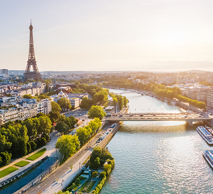 Vista panorâmica de Paris em um dia ensolarado. À esquerda, a Torre Eiffel se destaca no horizonte. O rio Sena atravessa o centro da imagem, com embarcações atracadas ao longo das margens. A cidade se estende dos dois lados do rio, com uma mistura de edifícios históricos e modernos. Árvores e vegetação verde acompanham as margens, trazendo contraste à paisagem urbana.