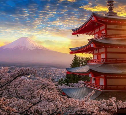 Paisagem do Japão com o Monte Fuji ao fundo, parcialmente coberto de neve. Em primeiro plano, há uma pagoda tradicional japonesa de cinco andares, pintada em vermelho e branco. Ao redor, cerejeiras em plena floração exibem flores rosa claro. O céu ao entardecer está colorido com tons de laranja e amarelo, criando uma atmosfera serena e encantadora. A imagem combina elementos naturais e culturais, representando a beleza típica do Japão.