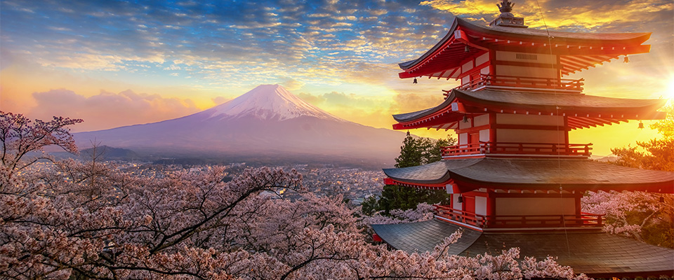 Paisagem do Japão com o Monte Fuji ao fundo, parcialmente coberto de neve. Em primeiro plano, há uma pagoda tradicional japonesa de cinco andares, pintada em vermelho e branco. Ao redor, cerejeiras em plena floração exibem flores rosa claro. O céu ao entardecer está colorido com tons de laranja e amarelo, criando uma atmosfera serena e encantadora. A imagem combina elementos naturais e culturais, representando a beleza típica do Japão.