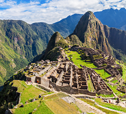 Vista panorâmica de Machu Picchu, antigo sítio arqueológico inca no Peru. A cidade está localizada no topo de uma montanha, com terraços verdes e ruínas de pedra bem preservadas. Ao fundo, montanhas altas cobertas por vegetação se destacam sob um céu azul parcialmente nublado. A imagem combina a grandiosidade das construções históricas com a beleza natural da paisagem montanhosa.