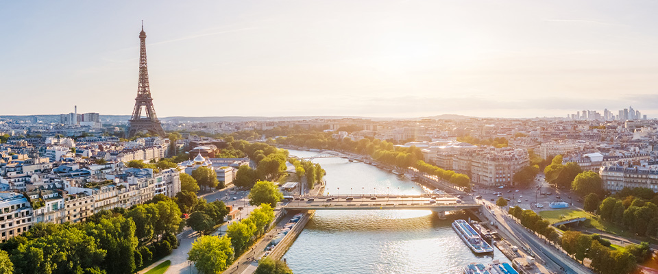 Vista panorâmica de Paris em um dia ensolarado. À esquerda, a Torre Eiffel se destaca no horizonte. O rio Sena atravessa o centro da imagem, com embarcações atracadas ao longo das margens. A cidade se estende dos dois lados do rio, com uma mistura de edifícios históricos e modernos. Árvores e vegetação verde acompanham as margens, trazendo contraste à paisagem urbana.