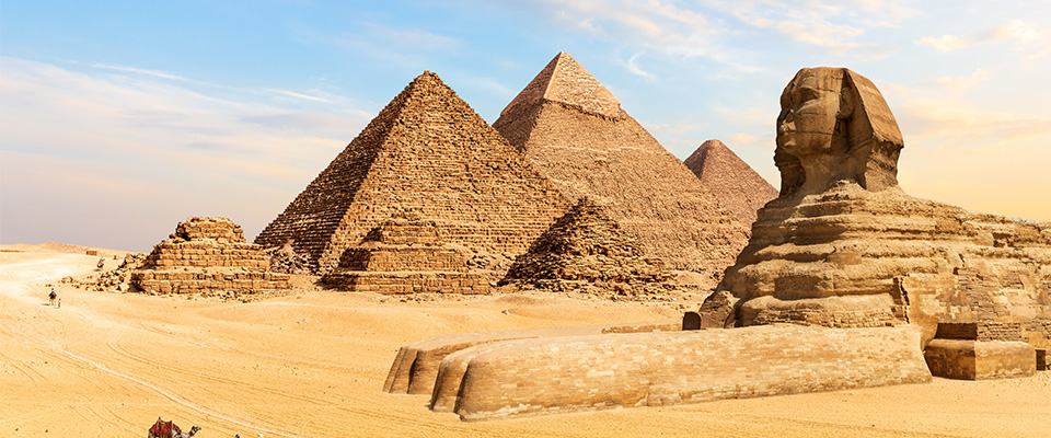 Imagem das Pirâmides de Gizé e da Grande Esfinge no Egito. No centro, três grandes pirâmides de pedra se destacam contra um céu azul com algumas nuvens. À direita, está a Grande Esfinge, uma escultura monumental com corpo de leão e cabeça humana. No canto inferior esquerdo, um camelo é conduzido por uma pessoa vestida com roupas típicas do deserto. O chão é coberto por areia dourada que se estende até o horizonte.