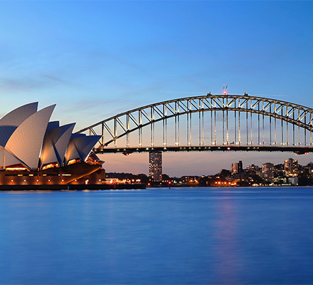 Vista panorâmica do Sydney Opera House e da Sydney Harbour Bridge ao entardecer. O céu exibe tons de azul e laranja típicos do pôr do sol. As luzes da cidade e dos edifícios estão acesas, refletindo na água calma do porto. À esquerda, o Sydney Opera House se destaca com seu design icônico em forma de velas brancas. Ao centro, a Sydney Harbour Bridge, uma ponte arqueada de aço, cruza a imagem. Ao fundo, arranha-céus iluminados compõem o horizonte da cidade de Sydney.