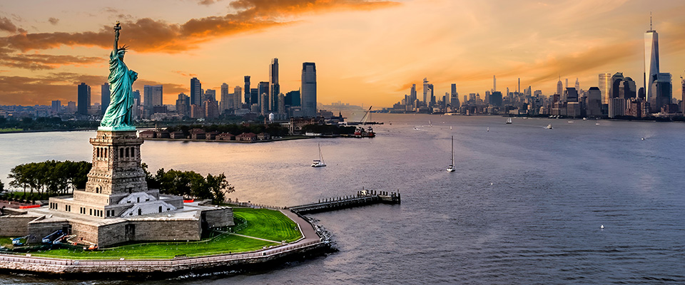 Imagem da Estátua da Liberdade em primeiro plano, situada na Liberty Island, cercada pelas águas do rio Hudson. Ao fundo, vê-se o horizonte da cidade de Nova York com diversos arranha-céus, incluindo o One World Trade Center à direita. O céu está parcialmente nublado, com tons alaranjados e rosados típicos do pôr do sol. Alguns barcos estão visíveis na água, compondo uma cena urbana e simbólica dos Estados Unidos.