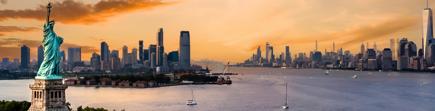 Imagem da Estátua da Liberdade em primeiro plano, situada na Liberty Island, cercada pelas águas do rio Hudson. Ao fundo, vê-se o horizonte da cidade de Nova York com diversos arranha-céus, incluindo o One World Trade Center à direita. O céu está parcialmente nublado, com tons alaranjados e rosados típicos do pôr do sol. Alguns barcos estão visíveis na água, compondo uma cena urbana e simbólica dos Estados Unidos.