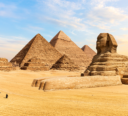 Imagem das Pirâmides de Gizé e da Grande Esfinge no Egito. No centro, três grandes pirâmides de pedra se destacam contra um céu azul com algumas nuvens. À direita, está a Grande Esfinge, uma escultura monumental com corpo de leão e cabeça humana. No canto inferior esquerdo, um camelo é conduzido por uma pessoa vestida com roupas típicas do deserto. O chão é coberto por areia dourada que se estende até o horizonte.
